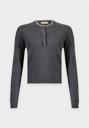 Femme portant un haut à manches longues gris foncé et un pantalon marron, bottes noires, ceinture noire, portant un sac à main marron foncé sur une épaule.
