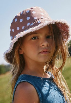 Noor tüdruk, kellel on pikad juuksed, pruuniga täpiline päikesemüts ja sinine varrukateta pluus, õues päikesevalguses.