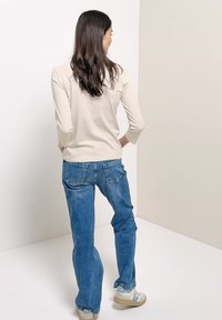 Femme aux longs cheveux foncés portant un haut beige clair, un jean bleu et des baskets blanches, debout dans une pièce aux couleurs neutres, vue de dos.