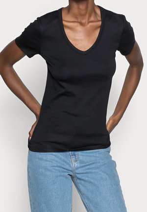 Vrouw die een zwart T-shirt met V-hals en korte mouwen en lichtblauwe jeans draagt, met handen in de zakken, tegen een witte achtergrond.