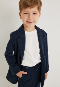 Navy blazer dengan kerah, lengan panjang, dan kantong, dipadukan dengan kaus putih polos dan celana navy dengan tali pengikat. Bahan halus, potongan kasual.
