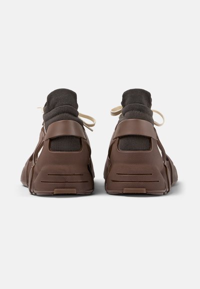 Sneaker marroni con una tomaia simile a una calza, caratterizzati da un tessuto testurizzato e dettagli in gomma modellata. Il design include sezioni ritagliate e lacci.