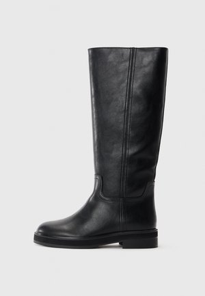 Bottes en cuir noir arrivant juste au genou, avec une texture lisse, un bout arrondi et un minimalisme dans les coutures. Semelle plate avec un petit talon.