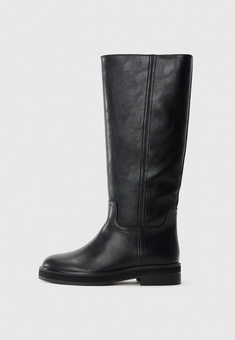 Bottes en cuir noir arrivant juste au genou, avec une texture lisse, un bout arrondi et un minimalisme dans les coutures. Semelle plate avec un petit talon.