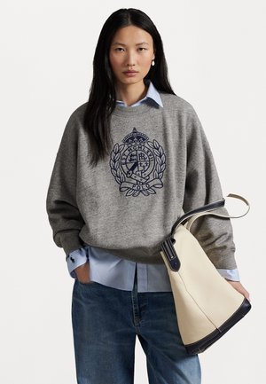 Grijze sweatshirt met marineblauwe logo, gedragen over een lichtblauw shirt. Blauwe denim jeans en een crème tas met donkere accenten maken de outfit compleet.
