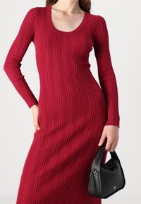 Robe rouge côtelée à manches longues avec un col rond. Le mannequin tient un sac à main en cuir noir de forme incurvée avec des détails en métal argenté.
