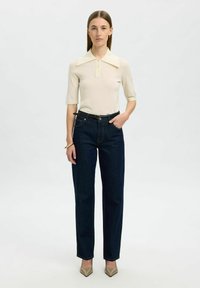 Camisa de polo de punto beige con un gran cuello, combinada con vaqueros de mezclilla oscuros de cintura alta, complementada con un cinturón negro y tacones de punta beige.