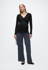 Έγκυος γυναίκα που φοράει μαύρο μακρυμάνικο τοπ, φαρδιά γκρι παντελόνια, μαύρα παπούτσια, κρατώντας ένα ασημί τσαντάκι μπροστά σε απλό φόντο.