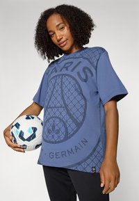 Blue cotton t-shirt with a large, geometric Paris Saint-Germain logo. Short sleeves, round neck, and a smooth texture. A soccer ball is held nearby.