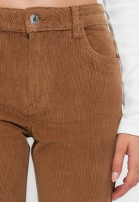 Brązowe spodnie kordurowe z pionowym prążkowanym wzorem, pięcioma kieszeniami, metalowym zapięciem na guzik w talii oraz lekkim streczem w tkaninie.
