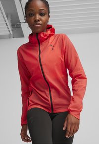 Leichte rote Jacke mit Kapuze, schwarzem Reißverschluss und frontalen Taschen, kombiniert mit dunklen Leggings, mit einem Logo auf der oberen linken Seite.