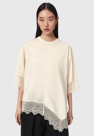 Vrouw draagt een losse crèmekleurige blouse met kanten afwerking aan de mouwen en zoom, staand tegen een effen lichtgrijze achtergrond.