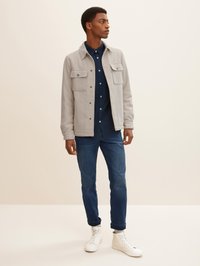 Graue Jacke mit Kragen und zwei Brusttaschen über einem blauen Hemd mit Knopf, kombiniert mit hochgekrempelten dunklen Jeans und weißen Sneakers mit hohem Schaft.