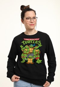 Černá mikina s barevným grafickým motivem čtyř kreslených želv. Design obsahuje tučný text "TEENAGE MUTANT NINJA TURTLES" nad postavami.