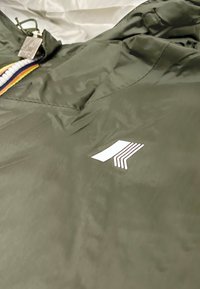 Giacca in nylon verde oliva con logo bianco e un tirazip multicolore. Texture liscia con leggero lucido.