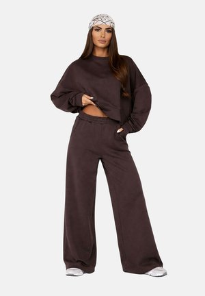 Femme aux longs cheveux portant un foulard blanc imprimé, un sweat-shirt oversize brun foncé, un pantalon à jambes larges et des baskets blanches, debout avec une main dans la poche.