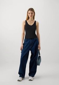 Sort ribstrikket bodystocking med V-udskæring, kombineret med løstsiddende mørkeblå denimjeans. Modellen holder en lille grå håndtaske. Hvide sneakers fuldender looket.