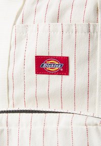 Șorț din țesătură bej cu dungi verticale roșii, prezentând un patch cu logo-ul Dickies în roșu și galben. Include o buzunar cu fermoar.