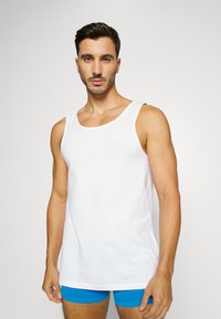 Débardeur blanc sans manches en coton avec un col rond et un petit logo en haut à gauche. Modèle portant un short bleu.