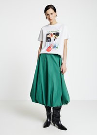 T-shirt graphique blanche avec imprimé vert et motif piscine, associée à une jupe verte fluide et des bottes noires à bout pointu.