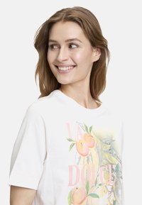 T-shirt en coton blanc à manches courtes, avec des graphiques colorés de pêches et de feuilles vertes et le texte "LA DOLCE" en rose.
