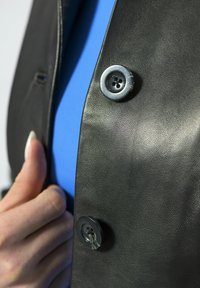 Detail van een zwart leren jack met een grote knop en een blauw shirt eronder. Textuur van leer met een gladde afwerking.