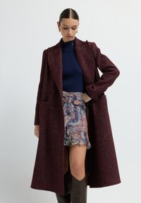 Abrigo de mezcla de lana burdeos con un patrón de espiga, diseño de doble botonadura y bolsillos laterales, combinado con un suéter azul y una falda con estampado.