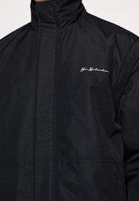 Svart jacka i slät tyg med hög krage. Har en vit broderad logotyp på bröstet och strukturerade sömdetaljer.