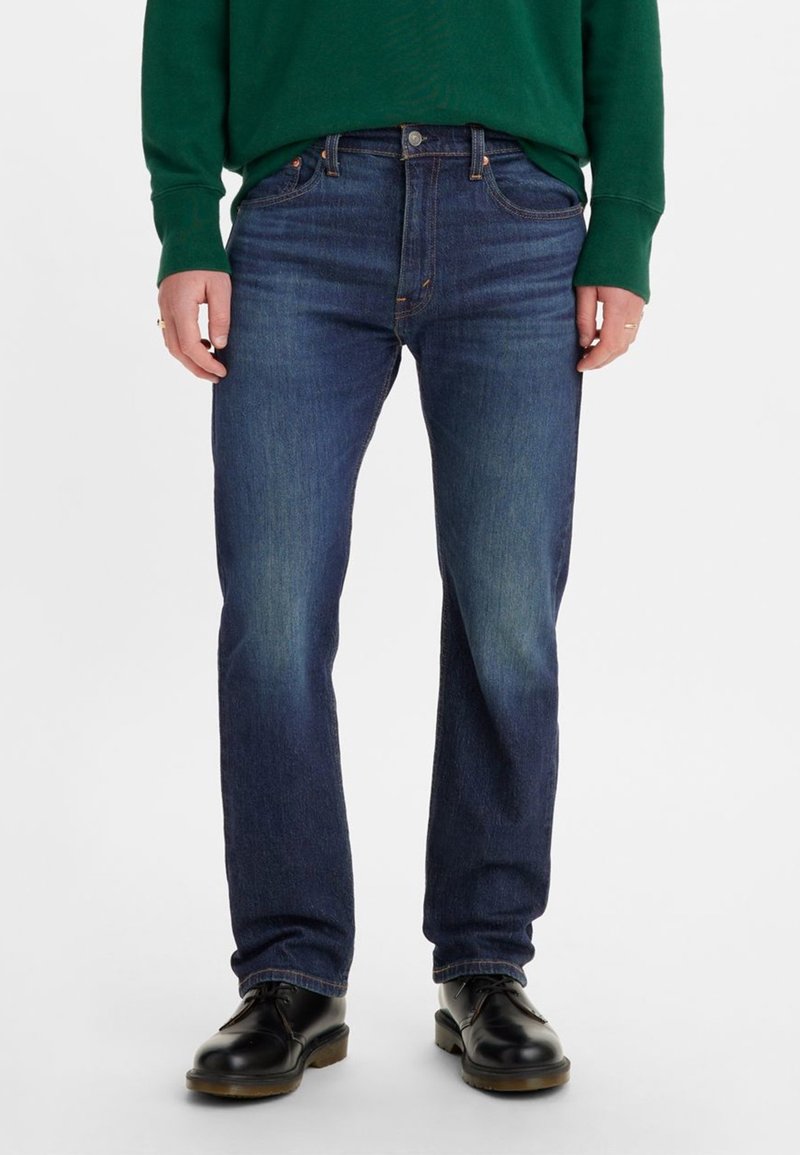 Homme portant un jean droit bleu foncé, des chaussures en cuir noir et un haut vert à manches longues, debout sur un fond blanc.