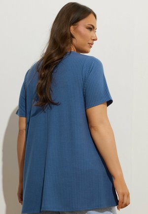 Vrouw met lang bruin haar die een losse, geribbelde, korte mouw blauwe top draagt, gezien van achteren tegen een effen lichte achtergrond.