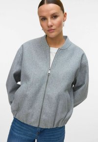Femme avec les cheveux raides tirés en arrière, portant des boucles d'oreilles pendantes, une veste zippée gris clair, un haut blanc et un jean bleu, devant un fond uni.