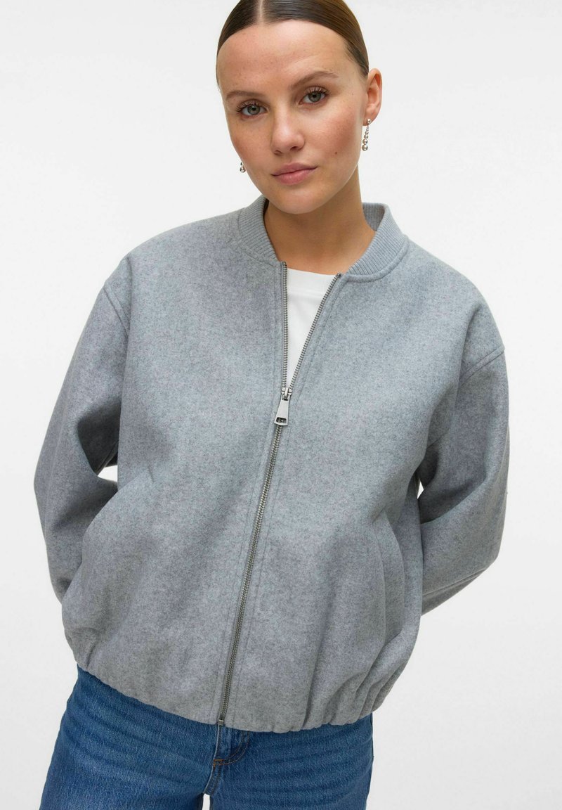 Femme avec les cheveux raides tirés en arrière, portant des boucles d'oreilles pendantes, une veste zippée gris clair, un haut blanc et un jean bleu, devant un fond uni.