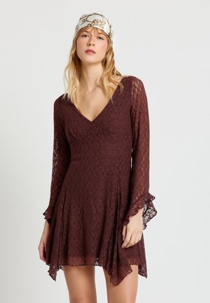 Femme blonde portant une robe longue en dentelle marron à manches longues avec un col en V et un foulard blanc à motifs, posant devant un fond uni.