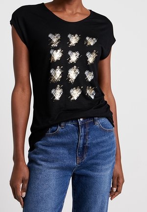 Zwart shirt met korte mouwen met 12 gouden, witte en zilveren abstracte hartvormen aan de voorkant, gedragen met blauwe spijkerbroek.