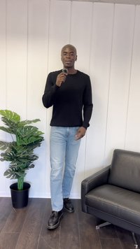 Black knit sweater, light blue jeans, black shoes; standing next to a potted green plant and a black leather sofa on wooden floor.