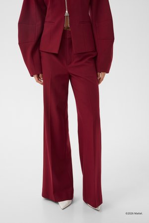 Άτομο που φοράει μπορντό παντελόνι ψηλόμεσο, με φαρδιά μπατζάκια και ταιριαστό σακάκι με φερμουάρ και φαρδιά μανίκια, συνδυασμένο με μυτερά λευκά παπούτσια.