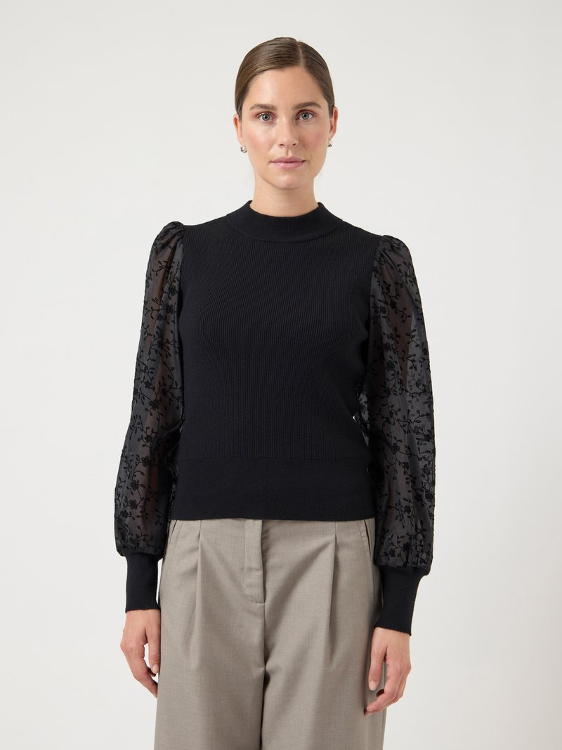 Black knitted sweater with sheer black floral-patterned sleeves and ribbed cuffs. Paired with light gray pleated trousers.