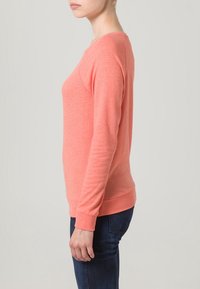 Korálově růžový top s dlouhým rukávem, volným střihem, kulatým výstřihem a žebrovanými manžetami, vyrobený z měkké látky. Kombinován s tmavě modrými džínami.