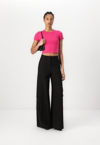 Zategnuti ružičasti crop top s kratkim rukavima i malim logom. Crne hlače širokih nogavica s džepovima. Model drži malu crnu torbicu.
