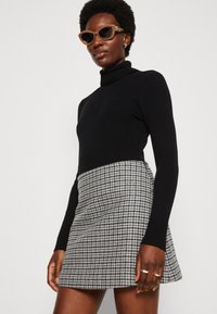 Schwarzer gerippter Rollkragenpullover kombiniert mit einem Rock im Hahnentrittmuster in Grau und Marineblau. Model trägt Sonnenbrillen und silberne Ringe.