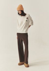 Biała oversize bluza z kieszenią i haftowanymi detalami, zestawiona z brązowymi spodniami o szerokich nogawkach i beżowymi wsuwanymi butami, uzupełniona pomarańczową czapką beanie.