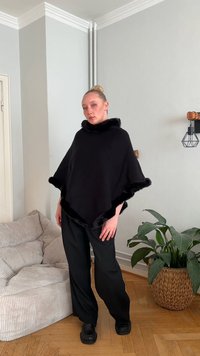 Svart poncho med pälsdetaljer, lös design och hög krage. Bärs över svarta vida byxor. Bakgrunden visar en texturerad vägg och en grön växt.