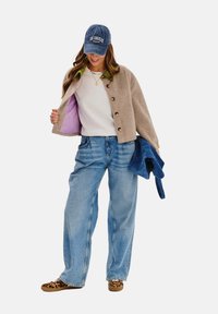 Beżowa kurtka z guzikami, fioletową podszewką, biała sweter, jasnoniebieskie luźne dżinsy i kapcie w panterkę, na głowie niebieska czapka.