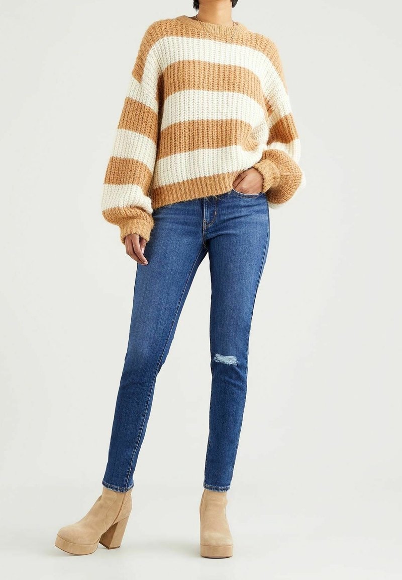 Personne portant un pull épais rayé beige et blanc, un jean slim bleu avec des genoux usés et des bottines beiges à talons, debout sur un fond clair.