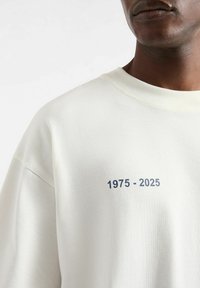 Sudadera de color crema claro con cuello redondo, que presenta una impresión numérica azul "1975 - 2025" en la parte frontal. Tejido suave y texturizado.
