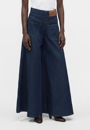 Jean bleu foncé à jambes larges avec poches avant et étiquette en cuir marron, porté avec un haut blanc côtelé et des chaussures marron à talons.