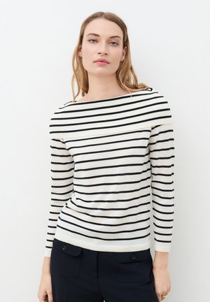 Jeune femme portant un pull blanc à rayures horizontales bleu marine et un pantalon noir, debout devant un fond blanc uni.