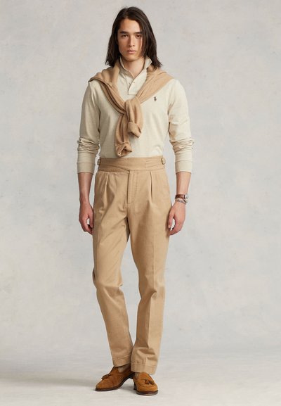 Beige long-sleeve polo with a light tan sweater tied over shoulders, beige pleated trousers, and brown suede loafers with tassels.