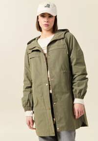 Veste légère vert olive avec capuche, fermeture à pression et zip, poignets élastiques et deux poches avant ; portée sur un pull crème.