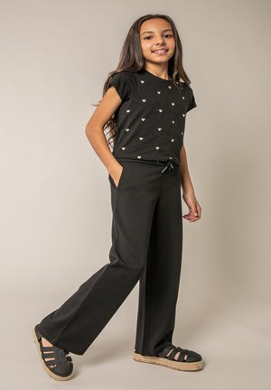 Fille aux longs cheveux portant un t-shirt noir avec de petits cœurs, un pantalon large noir et des sandales noires, debout avec un pied en avant et les mains dans les poches.